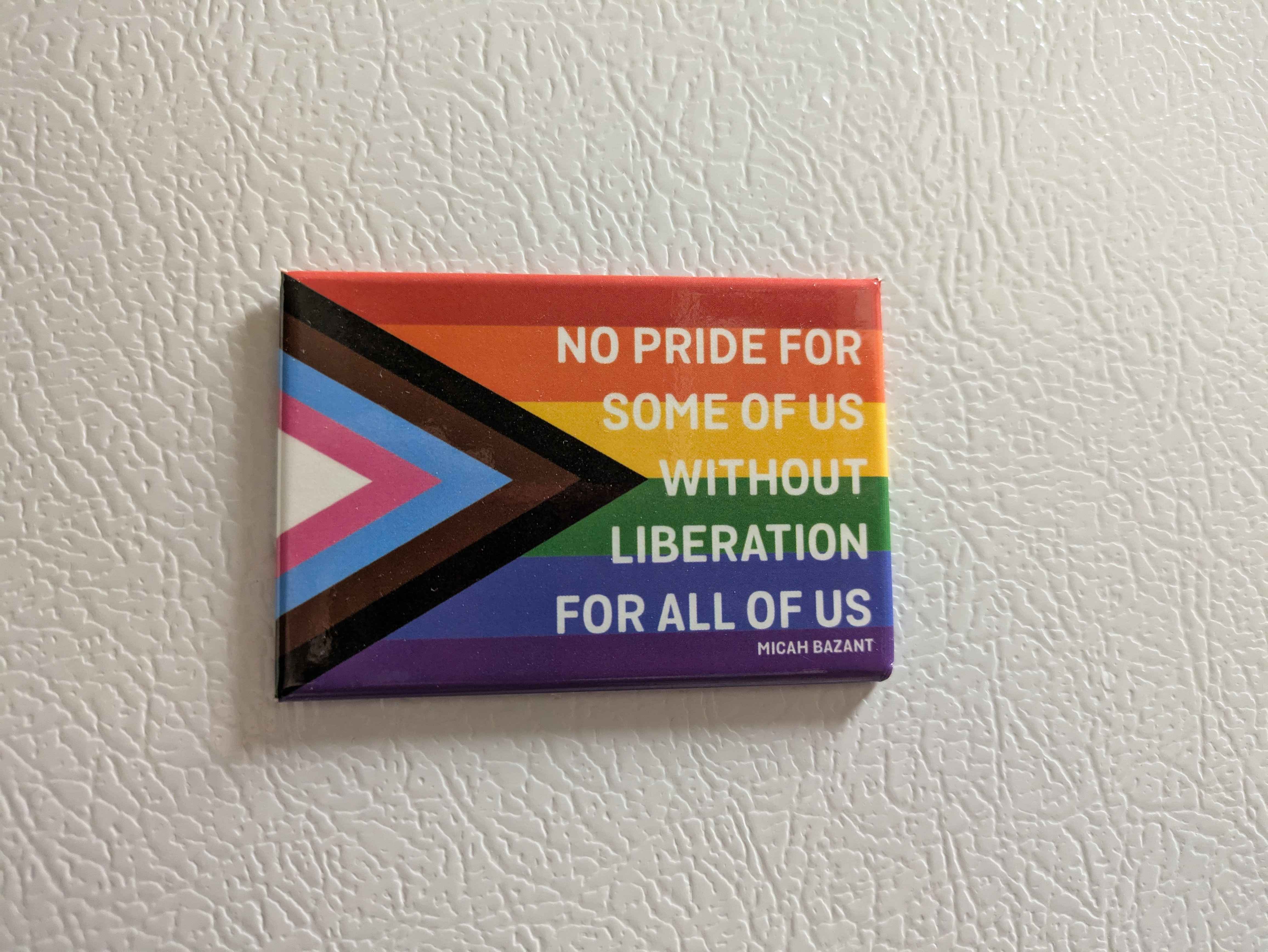 A picture of a magnet that has the progress pride flag on it with the quote 'No pride for some of us without liberation for all of us.' The magnet is on a white fridge.